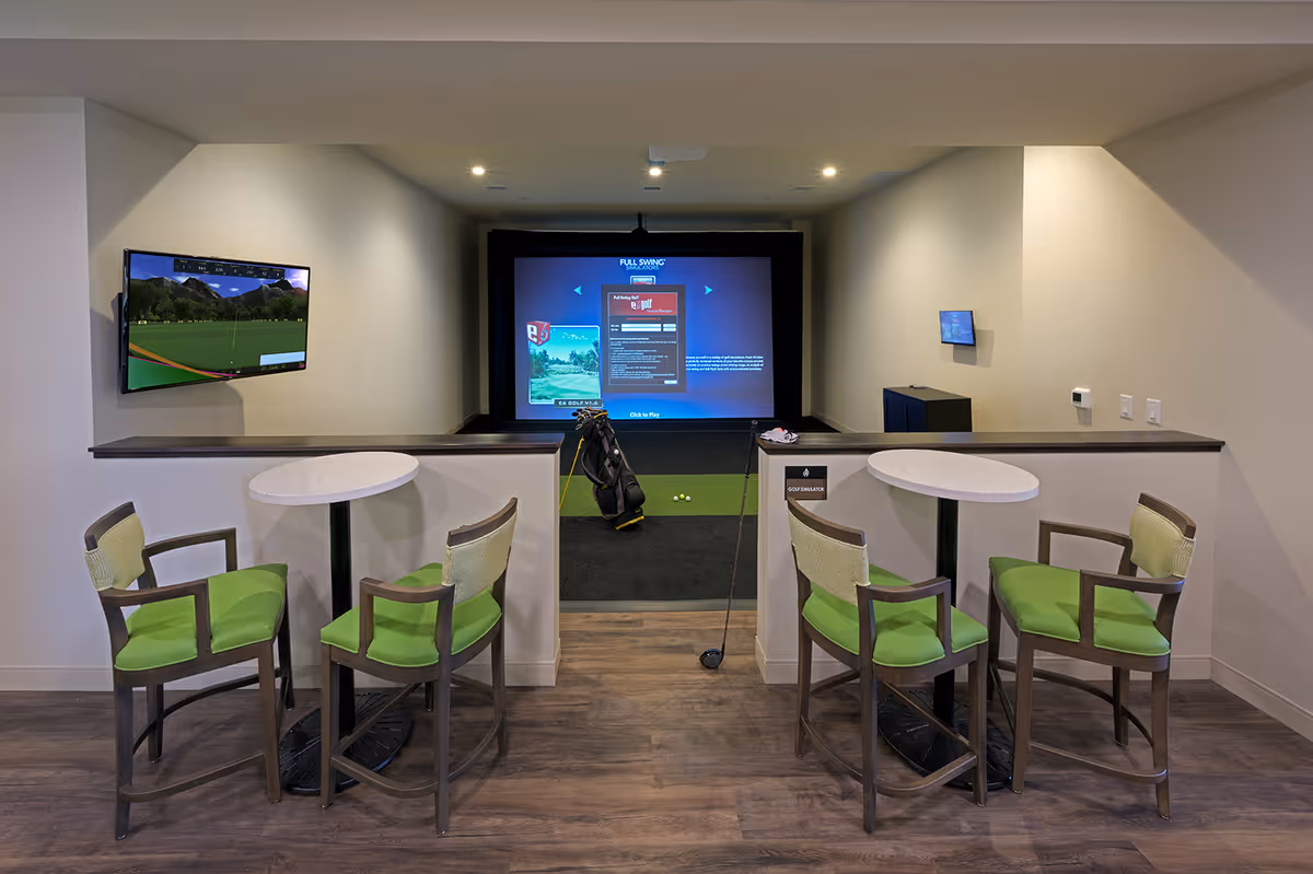Indoor recreational room with a golf simulator screen and bag, two round tables with green-cushioned chairs, and wall-mounted TVs.