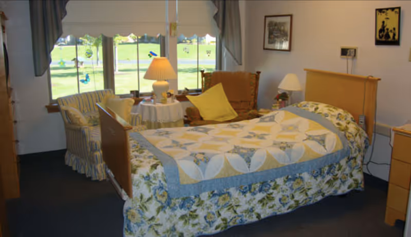 A cozy bedroom with a single bed covered in a floral and patchwork quilt. There is a wooden headboard and footboard. Next to the bed is a wooden nightstand with a lamp. In the corner near a window with white blinds and blue curtains, there is a small round table with a lamp and two chairs, one striped and one upholstered with a yellow pillow. The walls have framed pictures hanging.