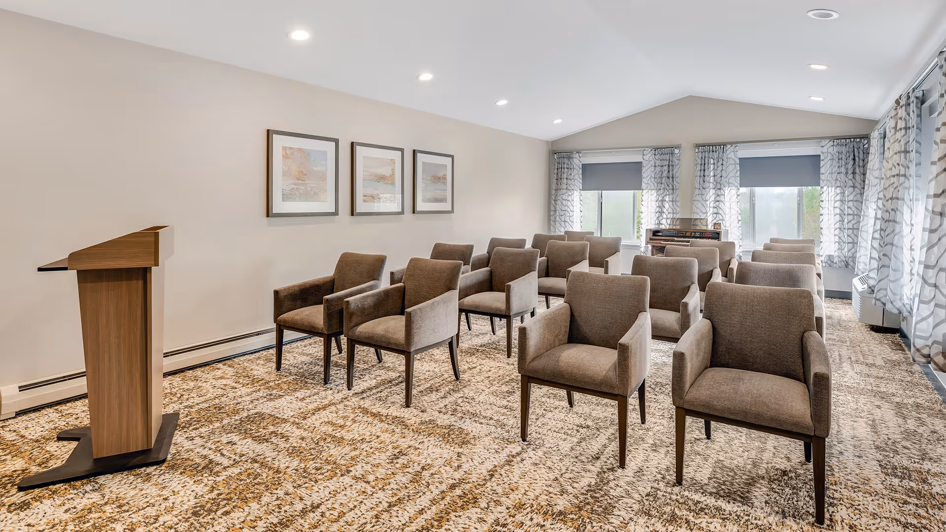 A small conference or meeting room with several rows of brown upholstered chairs arranged facing a wooden podium. The room has beige walls with three framed pictures, patterned carpet, and large windows with patterned curtains allowing natural light to enter.