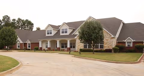 Exterior view of a single-story senior living facility building with a combination of brick and stone facade, multiple windows, a covered entrance with white columns, and a driveway in front. There are trees and well-maintained grass around the building.
