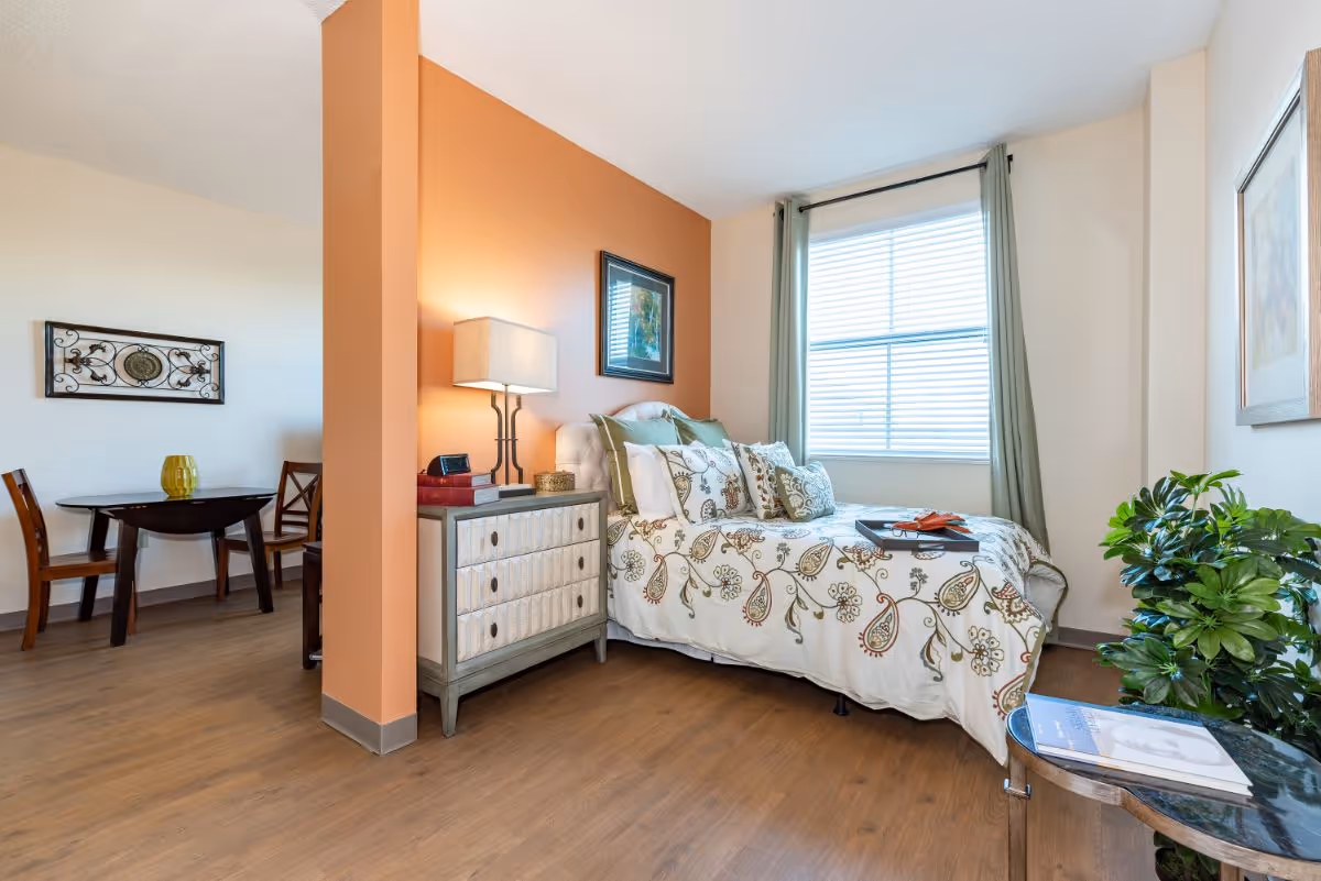 A bright bedroom with a bed covered in a floral patterned bedspread, multiple pillows, and a tray on top. Next to the bed is a nightstand with a lamp, books, and a clock. A large window with green curtains lets in natural light. To the left, there is a small dining area with a round table and two wooden chairs. The room has wooden flooring and light-colored walls, with one accent wall painted orange. A green plant and a small round table with a book are also visible.