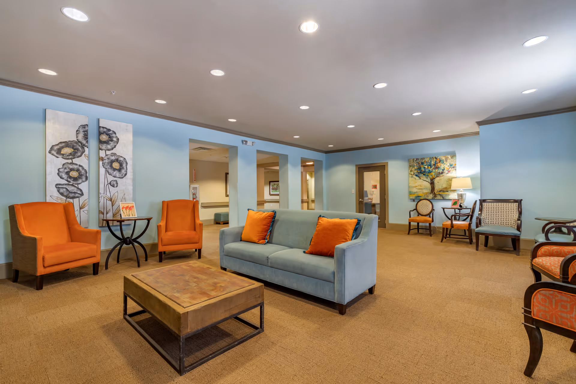 A spacious living room area with light blue walls and beige carpet. The room features a light blue sofa with orange pillows, two orange armchairs, a wooden coffee table, and several other chairs along the walls. There are two large floral paintings on one wall and a colorful tree painting on another. The ceiling has recessed lighting, and there is a table lamp on a side table near the back wall.