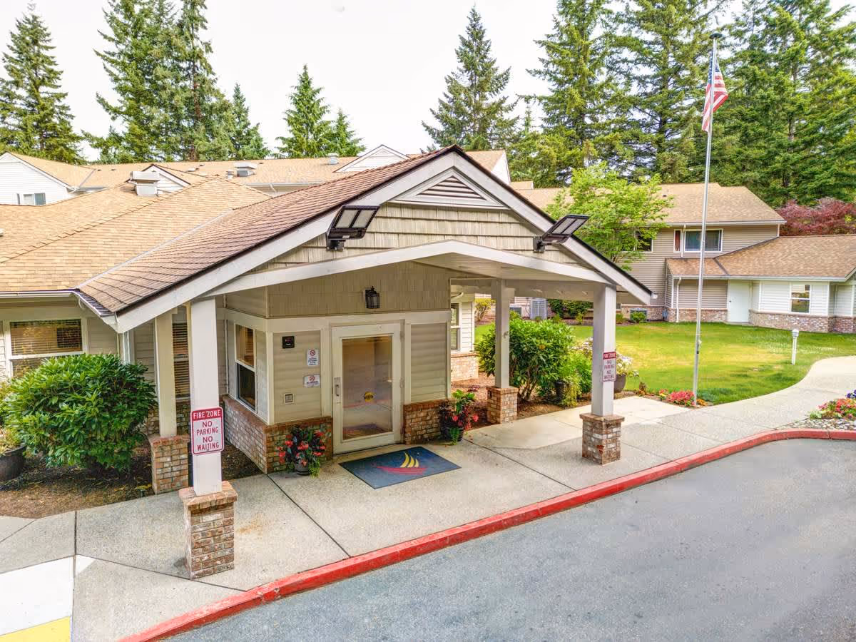 Entrance of a senior living facility with a covered drop-off area supported by brick pillars. The building has beige siding and a brown shingled roof. There are green bushes and flowers near the entrance, a flagpole with an American flag, and tall evergreen trees in the background.