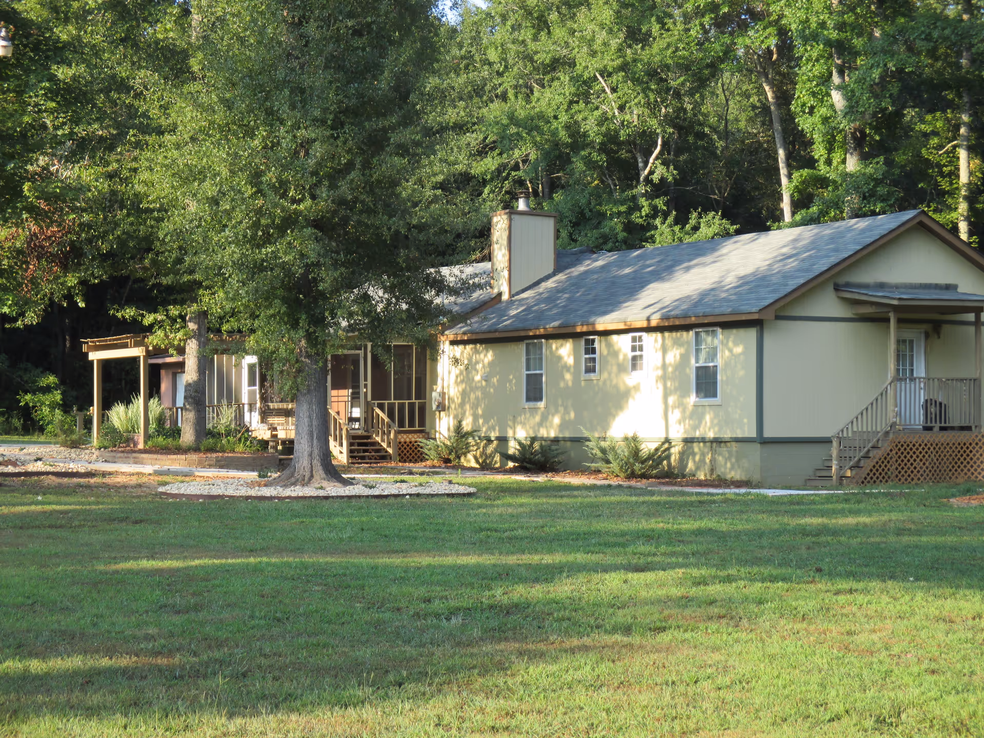 A single-story yellow building with a gray roof surrounded by green grass and trees. The building has several windows, a chimney, and two small porches with stairs leading up to them. The area appears to be peaceful and wooded.
