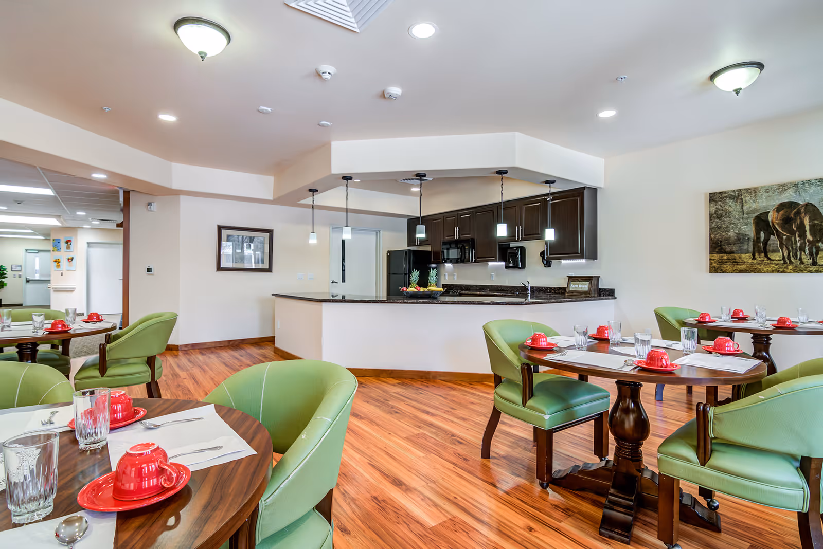 Dining area with round wooden tables set with red cups and saucers, glasses, and silverware. Green cushioned chairs surround the tables. In the background, there is a kitchen area with dark wood cabinets, a black refrigerator, microwave, and a countertop with pendant lights hanging above. The floor is wooden, and there is a painting of horses on the wall.