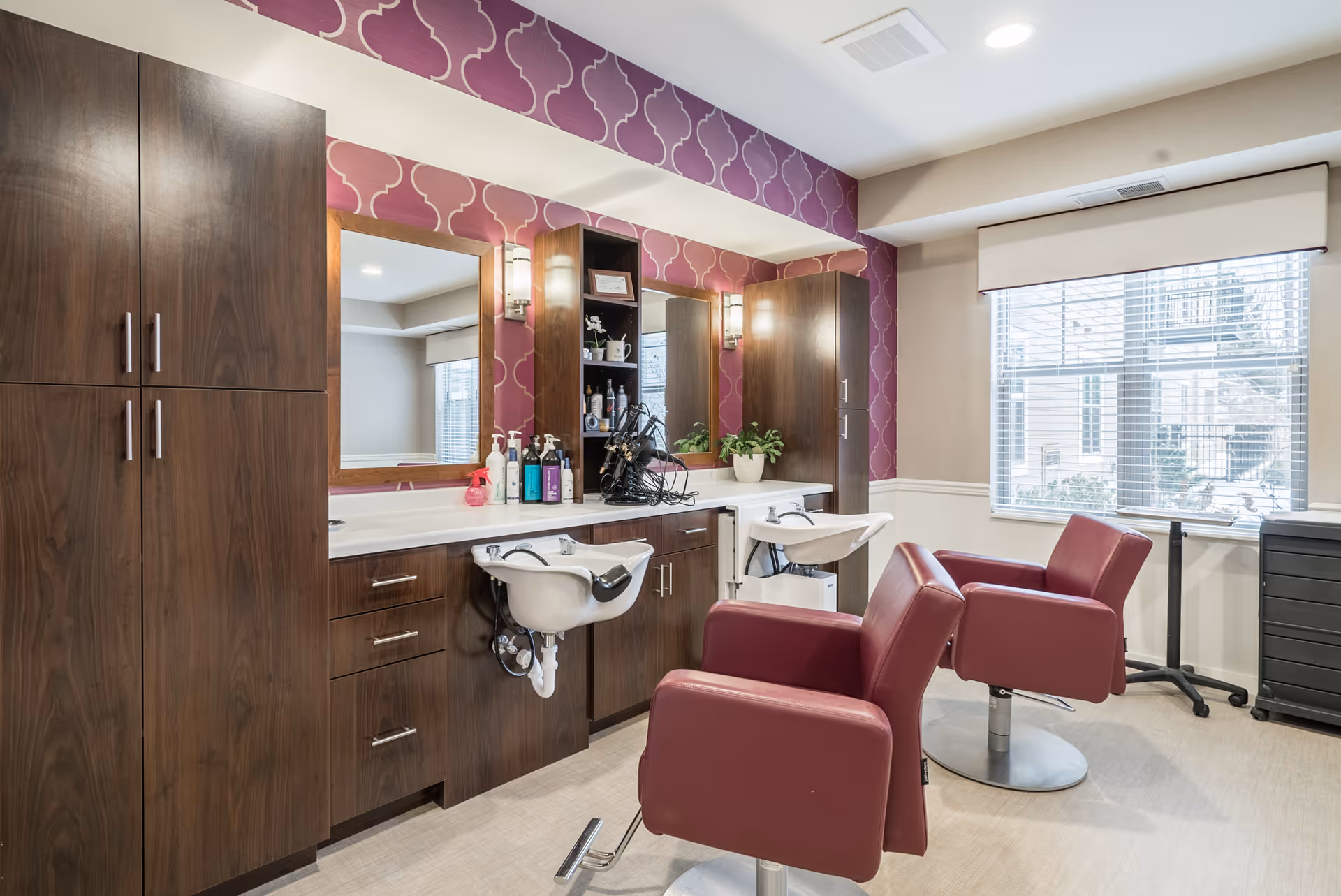 Interior view of a salon area in a senior living facility with two maroon salon chairs facing a counter with two sinks, mirrors, and wooden cabinets. The wall behind the counter has a purple patterned wallpaper, and a window with blinds lets in natural light.