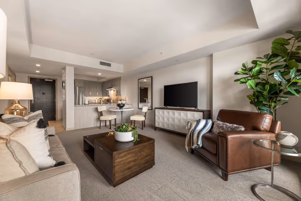 A modern living room with a beige sofa and a brown leather armchair, a wooden coffee table with a white planter on it, a flat-screen TV on a white geometric-patterned cabinet, a round glass side table with a decorative item, and a large green plant in the corner. In the background, there is a small dining area with a round table and two chairs, and a kitchen with light wood cabinetry and stainless steel appliances.