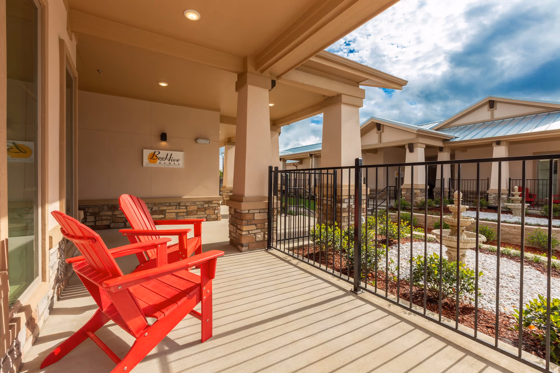 Outdoor patio area at BeeHive Homes of Frisco featuring two red Adirondack chairs on a concrete floor with a black metal fence overlooking a landscaped garden with a stone fountain and multiple building entrances under a partly cloudy sky.