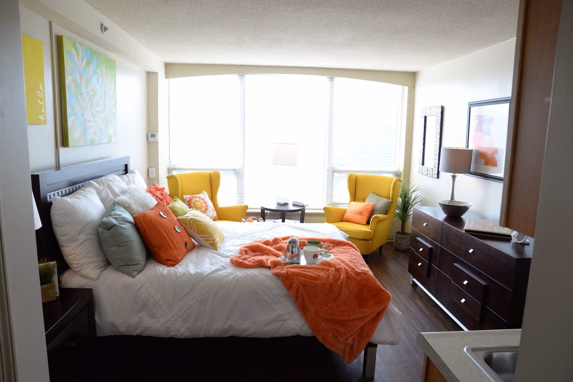 A bright bedroom with a large window covered by blinds letting in natural light. The room features a bed with white bedding and multiple colorful pillows, including orange and green ones. An orange throw blanket is draped over the bed with a tray holding a teapot and two cups. Two yellow armchairs with decorative pillows are positioned near the window with a small round table and a lamp between them. A dark wooden dresser with a lamp, a framed picture, and a mirror hangs on the wall. The floor is dark wood, and there is a small kitchenette sink visible in the foreground.