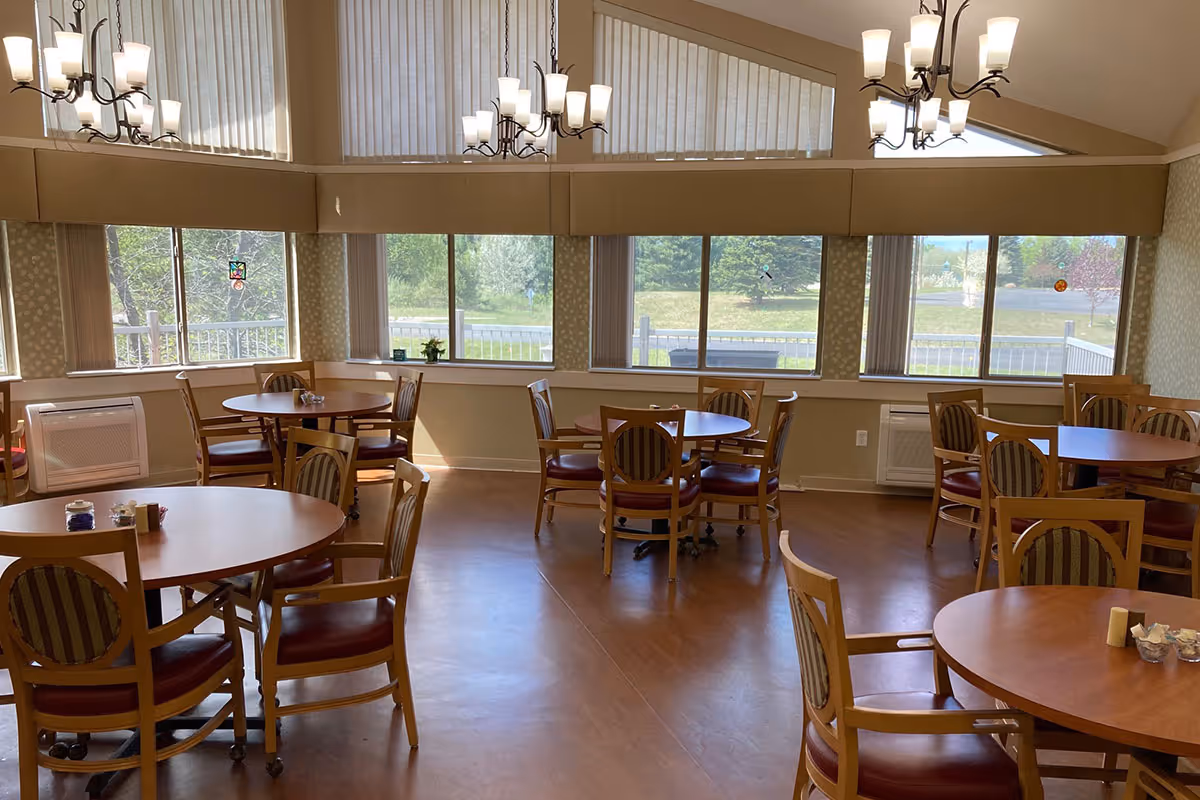 A bright dining room with multiple round wooden tables and chairs arranged neatly. Large windows with vertical blinds allow natural light to fill the room, and two chandeliers hang from the ceiling. Outside, greenery and trees are visible through the windows.