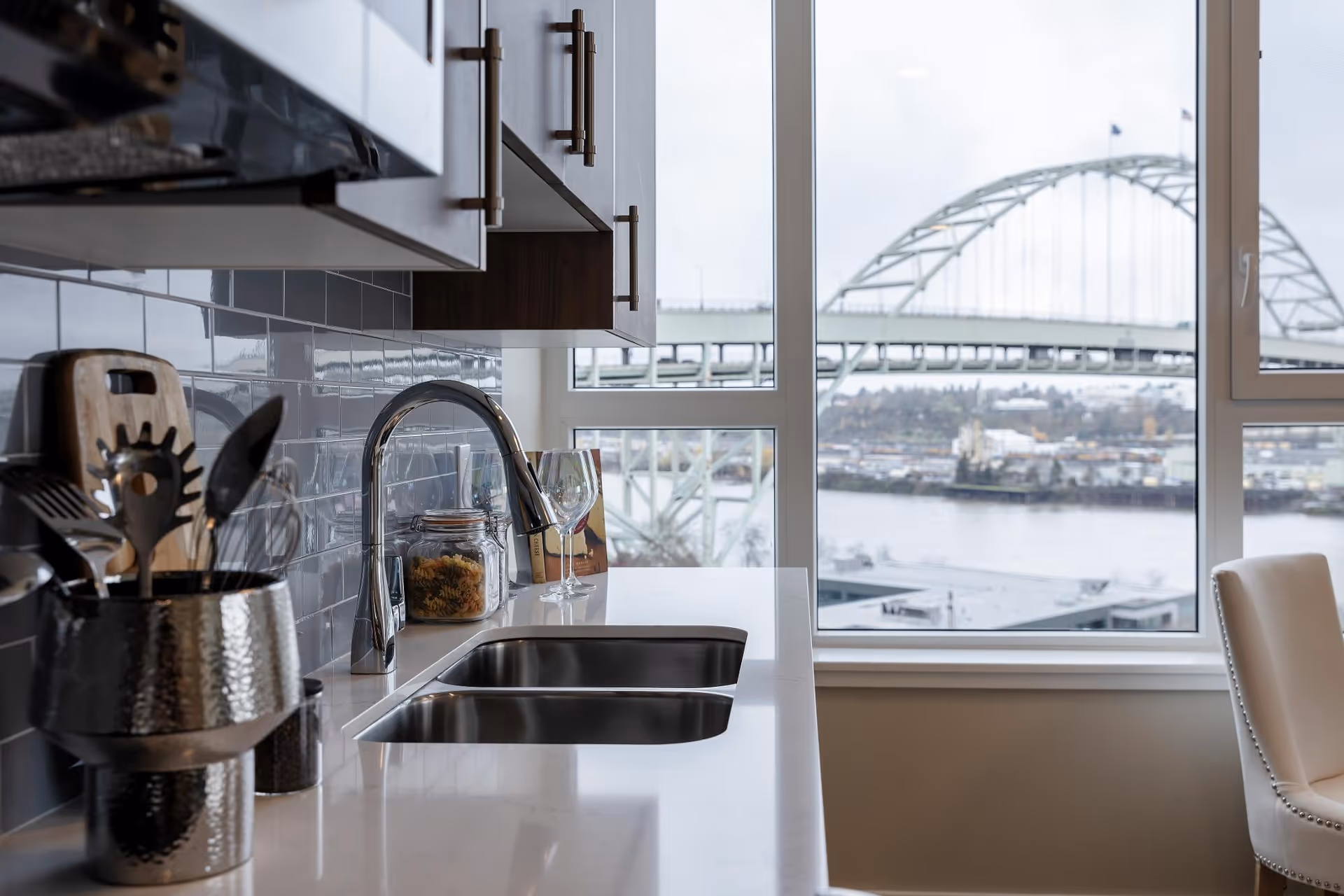 Modern kitchen countertop and sink with utensils and glassware, a large window showing a river and arched bridge outside.