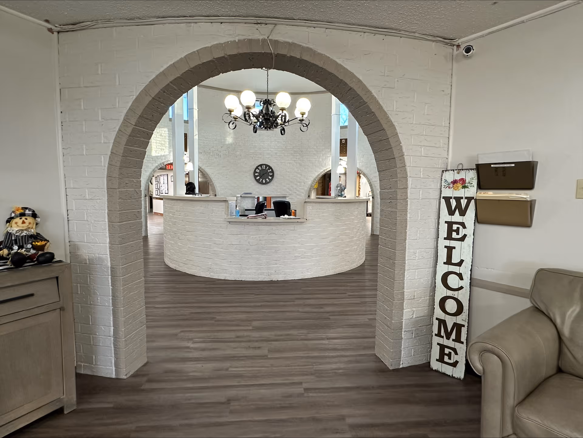 Interior view of a nursing and rehab facility reception area seen through a white brick archway. The reception desk is curved and white brick with two black chairs behind it. A black chandelier with lit bulbs hangs from the ceiling. A clock is mounted on the white brick wall behind the desk. To the right, there is a vertical wooden sign with the word 'WELCOME' and a beige leather armchair. To the left, a small cabinet with a decorative scarecrow figure is visible.