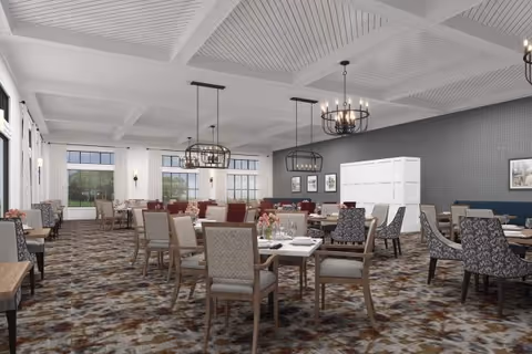A spacious dining room with multiple tables and chairs arranged neatly. The room features large windows allowing natural light to fill the space, decorative chandeliers hanging from a white coffered ceiling, and patterned carpet flooring. The walls are painted in neutral tones with some framed artwork and a white partition visible in the background.
