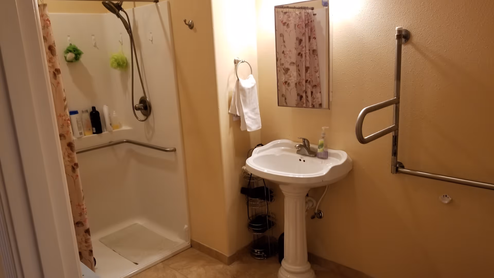 A bathroom with a walk-in shower featuring a handheld showerhead and grab bars. There is a pedestal sink with a soap dispenser, a small mirror above it, a towel ring with a white towel, and a floral shower curtain partially visible. The walls are painted beige and the floor is tiled.