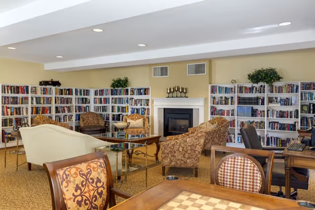 A cozy library or common area with multiple bookshelves filled with books along the walls. The room features a fireplace in the center, surrounded by various upholstered chairs and sofas arranged for conversation. There is a wooden table with a chessboard pattern in the foreground and a computer desk with a chair on the right side. The walls are painted light yellow and the ceiling has recessed lighting.