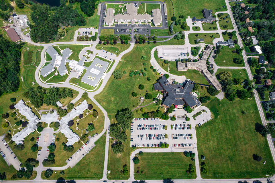 Aerial view of the Brothers of Mercy senior living facility showing multiple buildings, parking lots filled with cars, green lawns, trees, and surrounding roads.