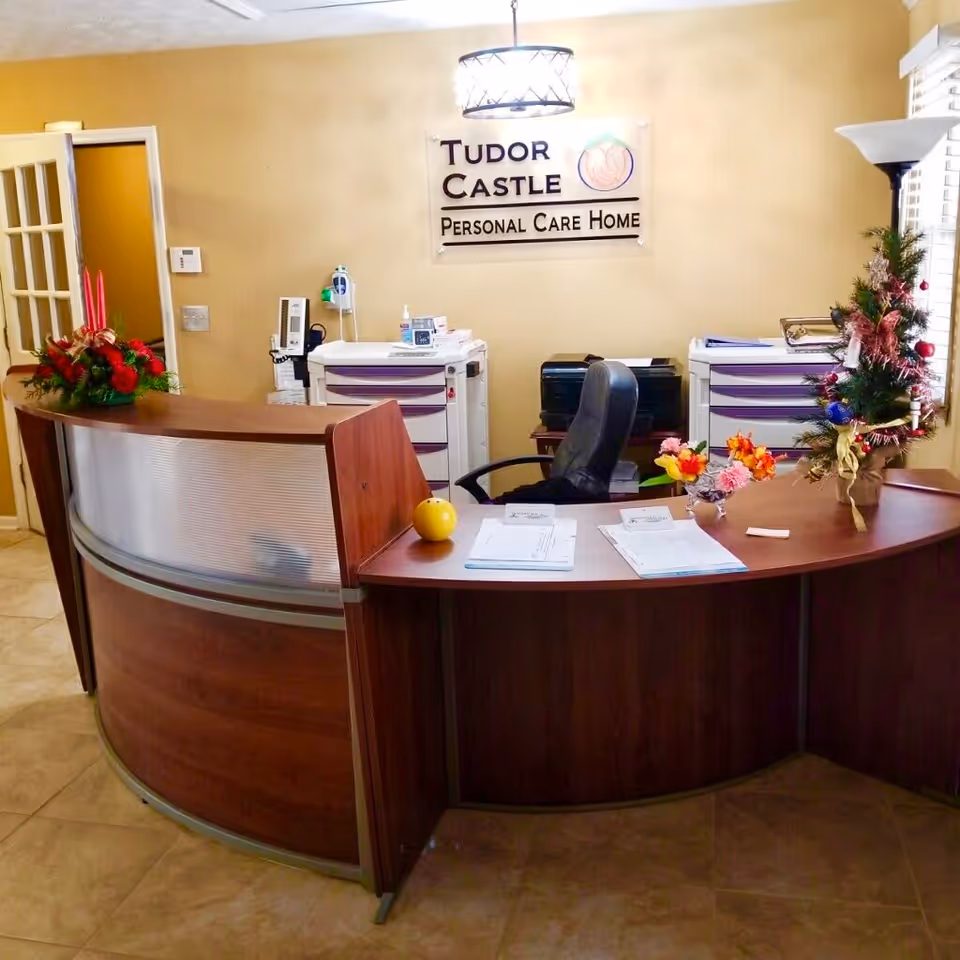 Reception area of Tudor Castle Personal Care Home featuring a curved wooden desk with paperwork, a black office chair, floral arrangements, and medical equipment against a beige wall with a sign displaying the facility's name.