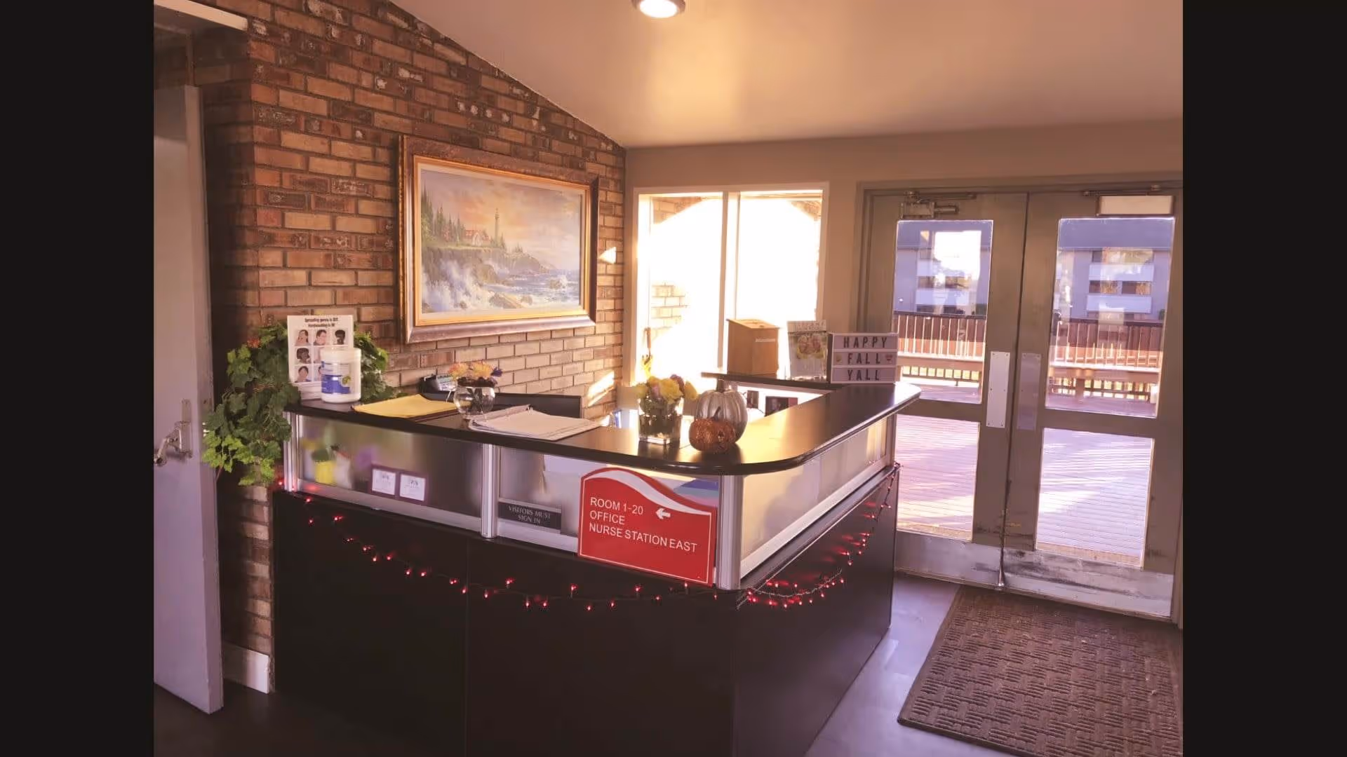 Reception desk inside a senior living facility with a brick accent wall, framed painting, front doors, and seasonal decorations.