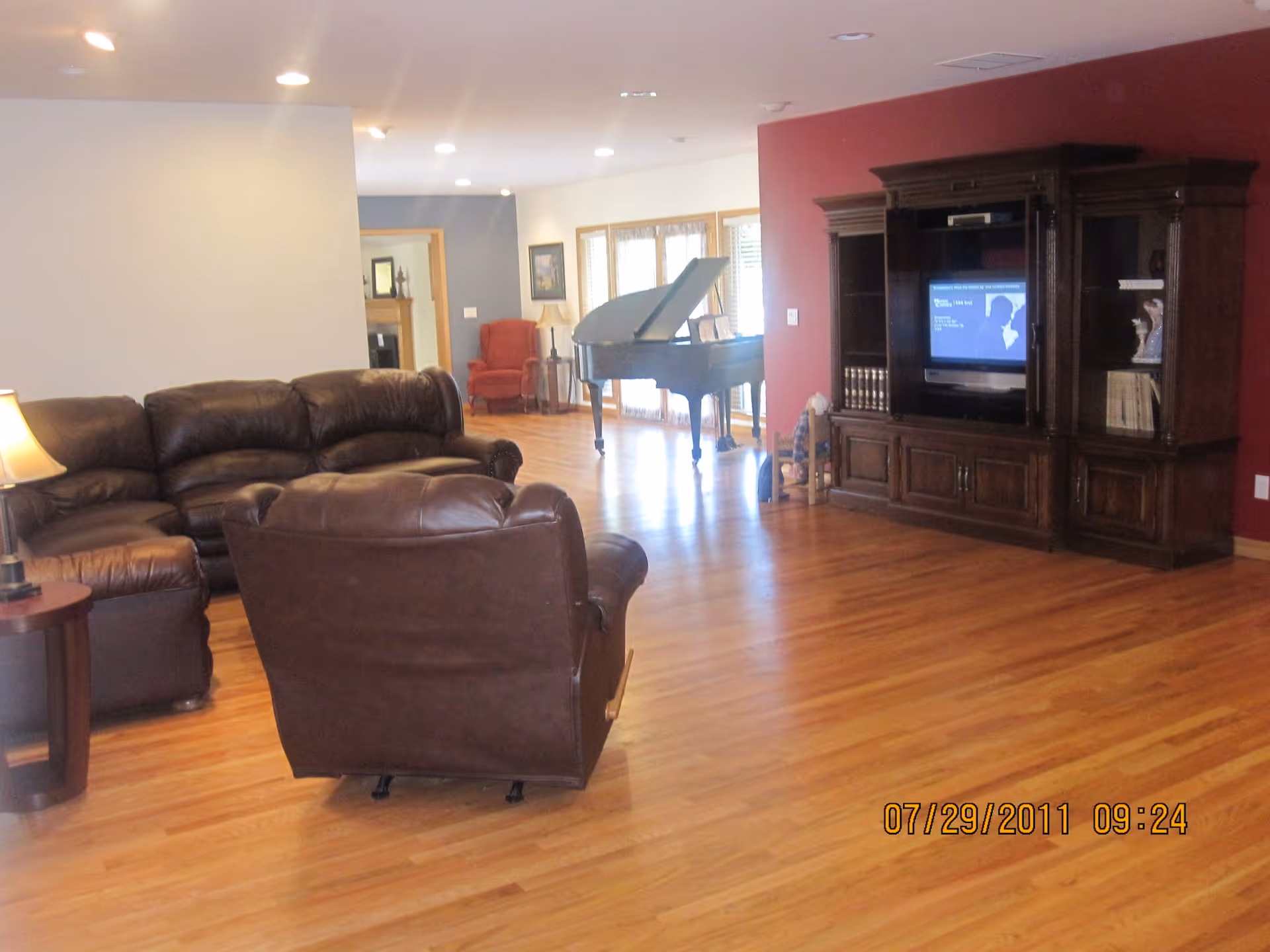 A spacious living room with wooden flooring featuring a brown leather sectional sofa and a matching armchair. There is a wooden entertainment center with a TV on a red accent wall. In the background, a black grand piano is visible near large windows letting in natural light. The room has recessed ceiling lights and a small side table with a lamp next to the sofa.