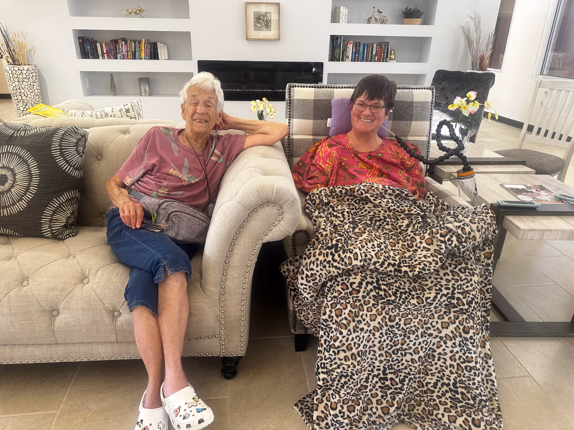 Two older women smiling while seated in a bright assisted-living common area—one on a sofa and one in a recliner covered with a leopard-print blanket.