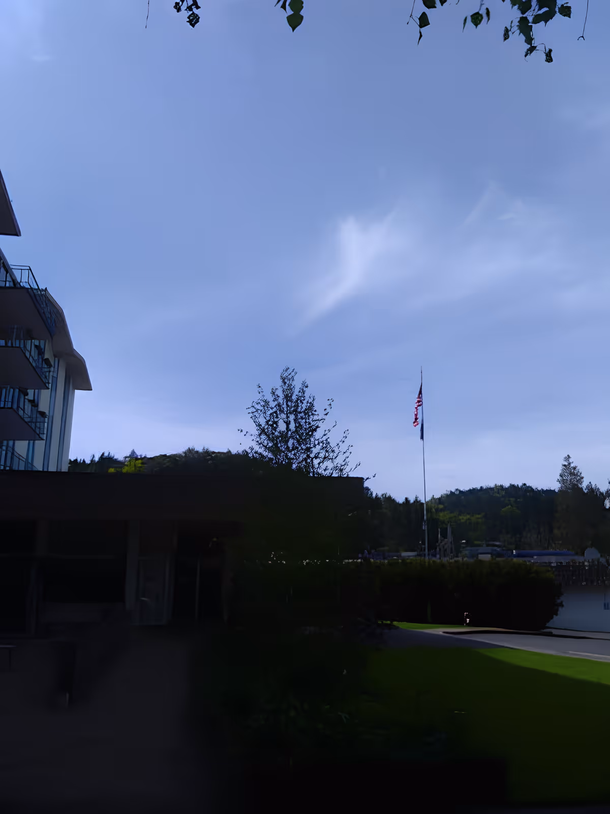 Outdoor view of a senior living facility with a multi-story building on the left, a flagpole with an American flag in the center, trees, bushes, and a clear blue sky with some clouds.