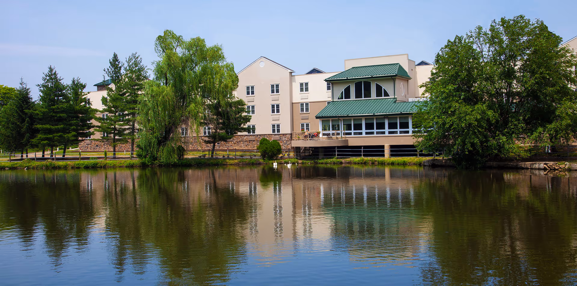 View of Commonwealth Senior Living At Willow Grove building with a green roof, surrounded by trees and reflected in a calm pond in the foreground under a clear blue sky.