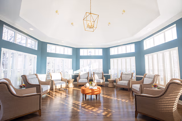 A bright and spacious sitting area with multiple wicker armchairs arranged in a circle around a round orange ottoman. The room features large windows with white shutters, teal walls, and a high white ceiling with a modern gold chandelier. The wooden floor reflects natural light coming through the windows.