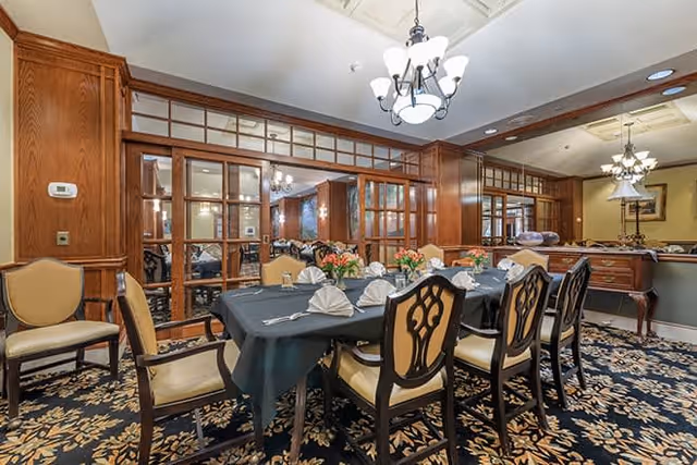 A formal dining room with a long table covered by a dark tablecloth, set with folded white napkins, silverware, and floral centerpieces. The room features wooden paneling, large mirrors, and elegant chandeliers hanging from the ceiling. Upholstered chairs surround the table, and the carpet has a floral pattern.