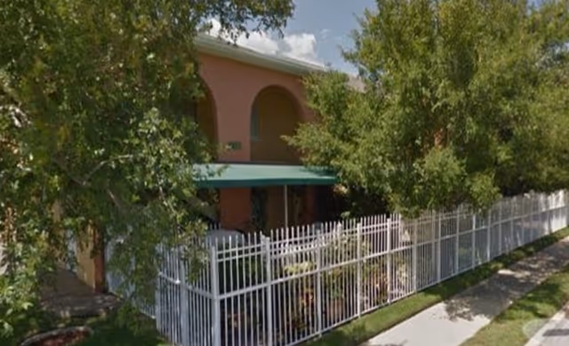 Exterior view of a two-story building with a peach-colored facade partially obscured by trees. The building has an arched window and a green awning over the entrance. A white metal fence surrounds the property, and a sidewalk runs alongside the fence.