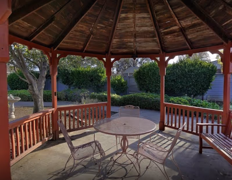 Wooden gazebo with a round metal table, three metal chairs and a bench overlooking landscaped bushes and trees.