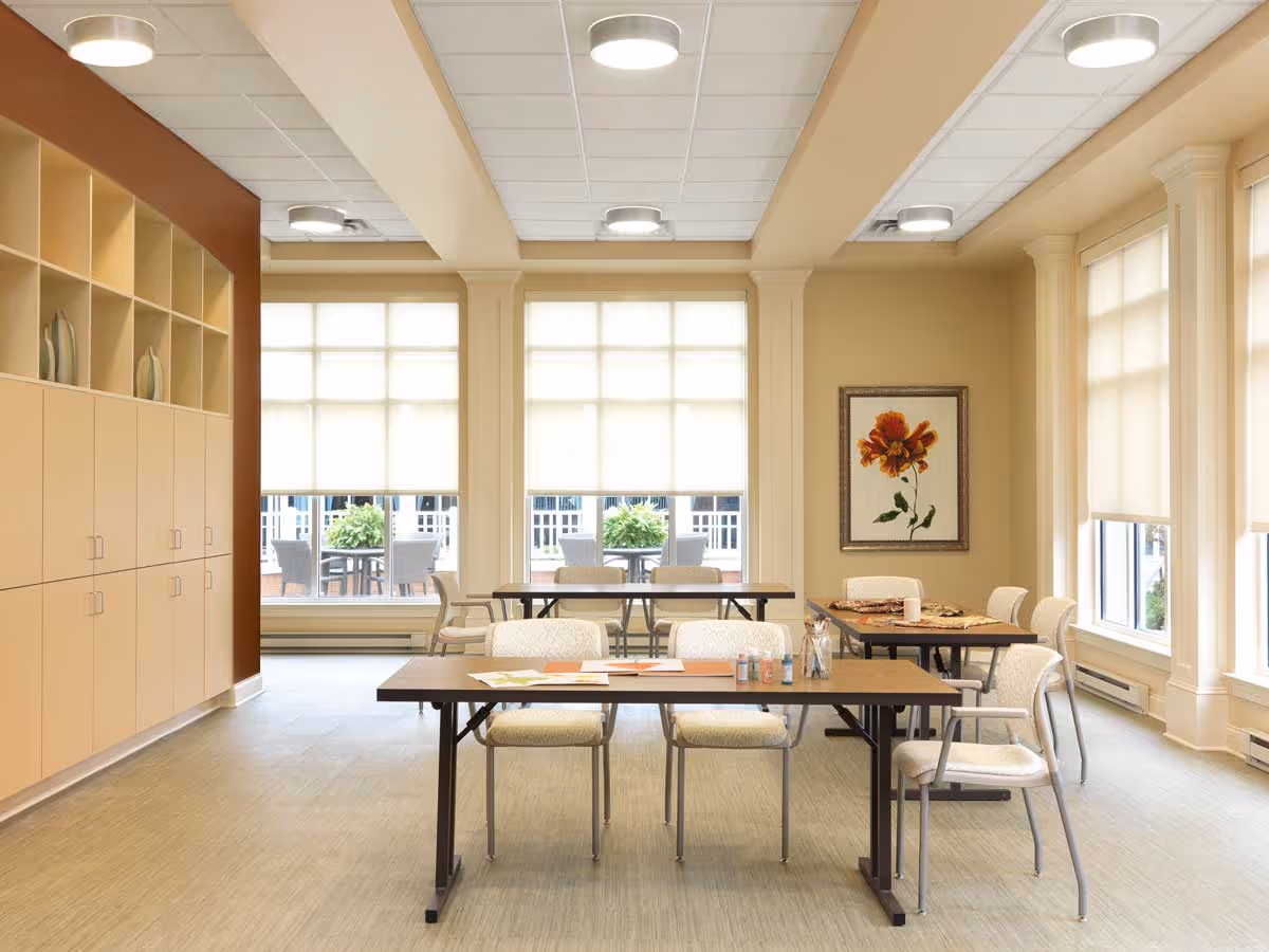 A bright, spacious room with large windows covered by white blinds, beige walls, and ceiling lights. The room contains several tables and chairs arranged for activities, with art supplies and papers on the front table. There is a framed picture of a flower on the wall and built-in cabinets on the left side.