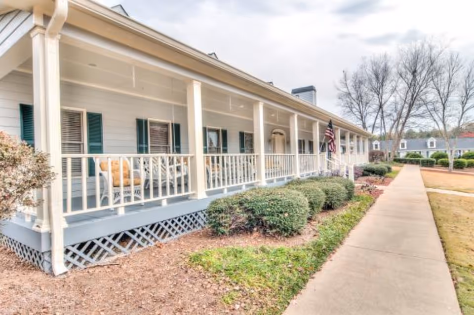 Long covered front porch of a single-story building with white railings, rocking chairs, an American flag, and a sidewalk lined with shrubs.