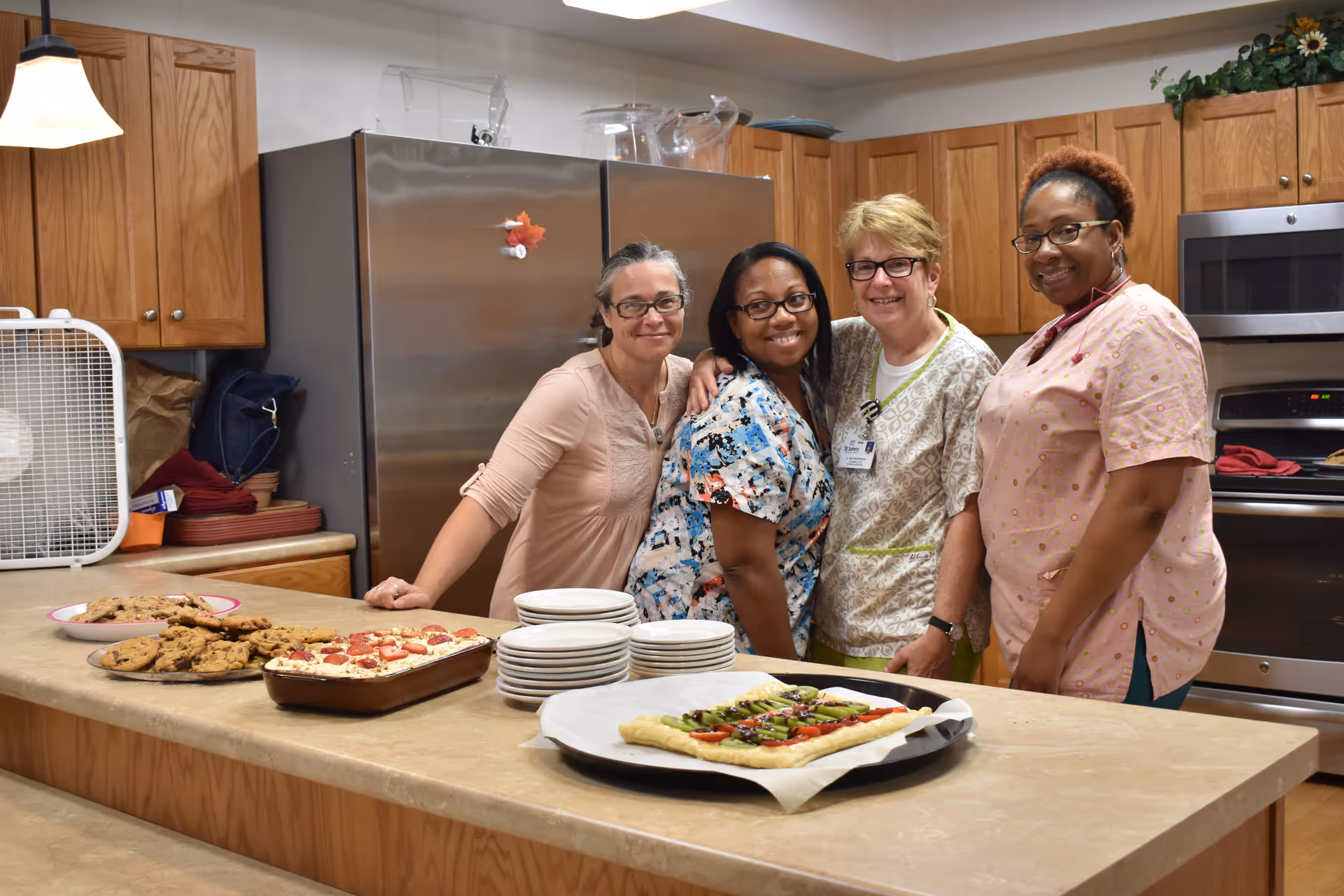 Four women standing together and smiling in a kitchen with wooden cabinets and stainless steel appliances. On the counter in front of them are plates of cookies, a dish of food topped with pepperoni, and a rectangular pizza with vegetables.