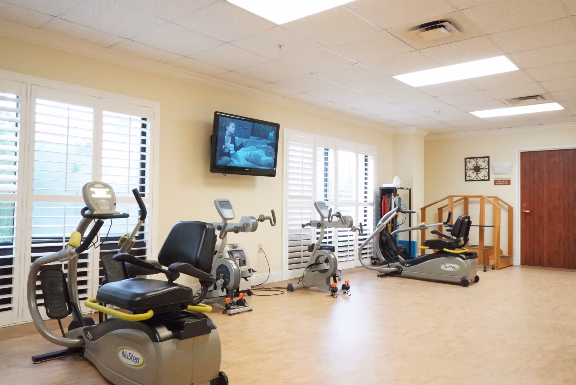 A well-lit exercise room with several pieces of fitness equipment including recumbent bikes and arm exercise machines. The room has large windows with white shutters, a wall-mounted TV showing a black and white program, and a wooden handrail structure near a closed wooden door.