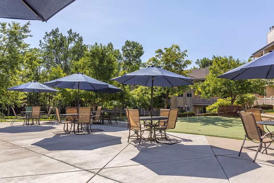 Outdoor patio area with multiple round tables and chairs, each shaded by large blue umbrellas. The patio is surrounded by green trees and grass, with a building visible in the background under a clear blue sky.