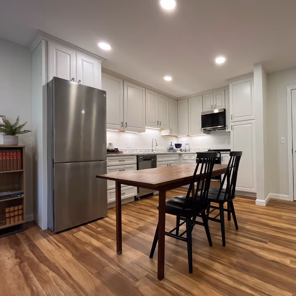 A modern kitchen with white cabinetry, stainless steel refrigerator, built-in microwave, dishwasher, and a wooden table with two black chairs on a wood floor. The kitchen is well-lit with recessed ceiling lights.