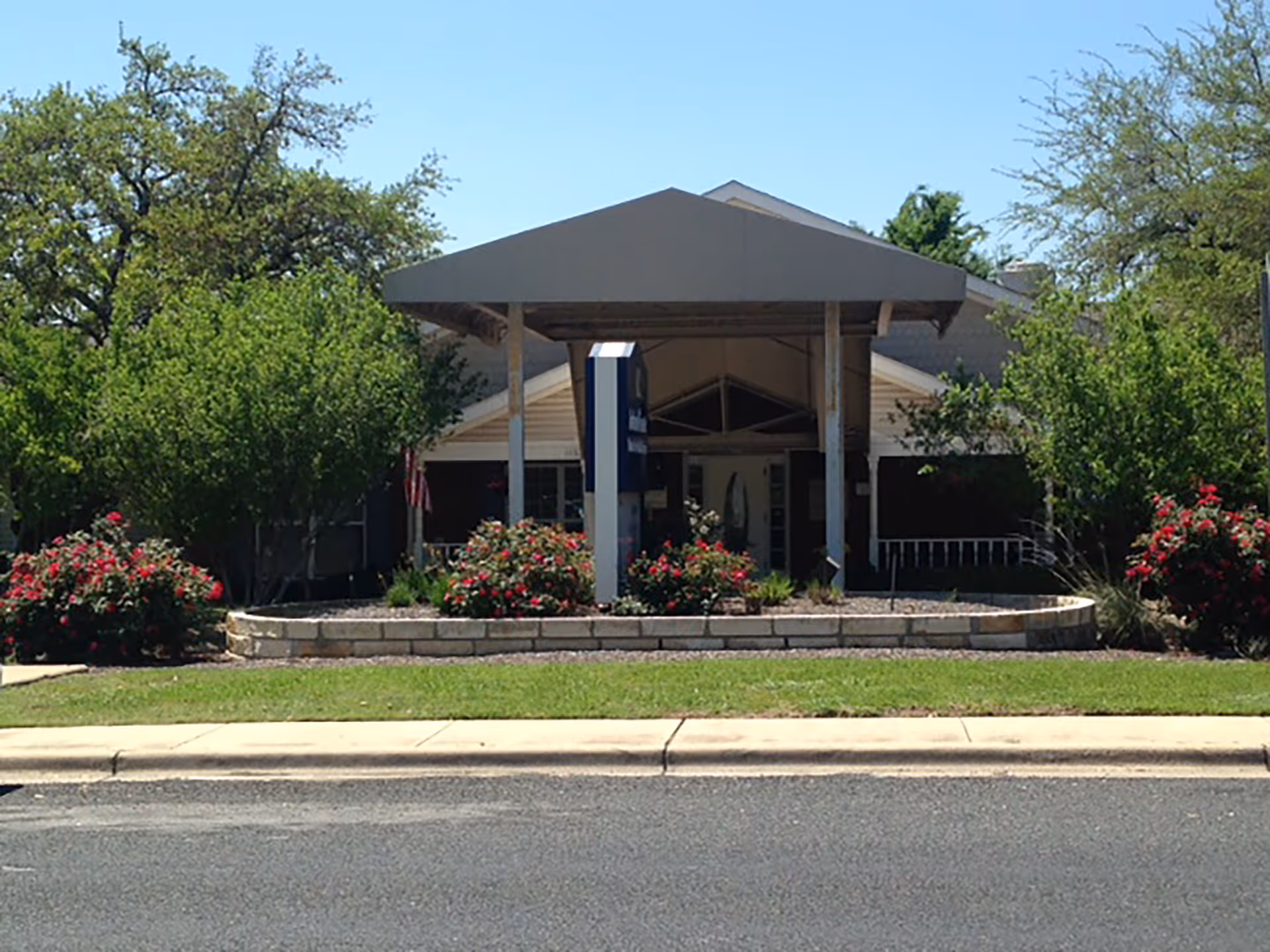 Front exterior view of Arden Courts - ProMedica Memory Care Community in Austin, showing the entrance with a covered canopy, surrounded by green trees and bushes with red flowers under a clear blue sky.