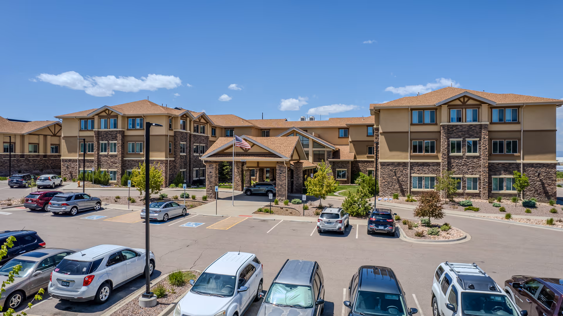 Exterior view of Pine Grove Crossing senior living facility showing a large three-story building with stone and beige siding, a covered entrance with an American flag, and a parking lot with several cars under a blue sky with scattered clouds.