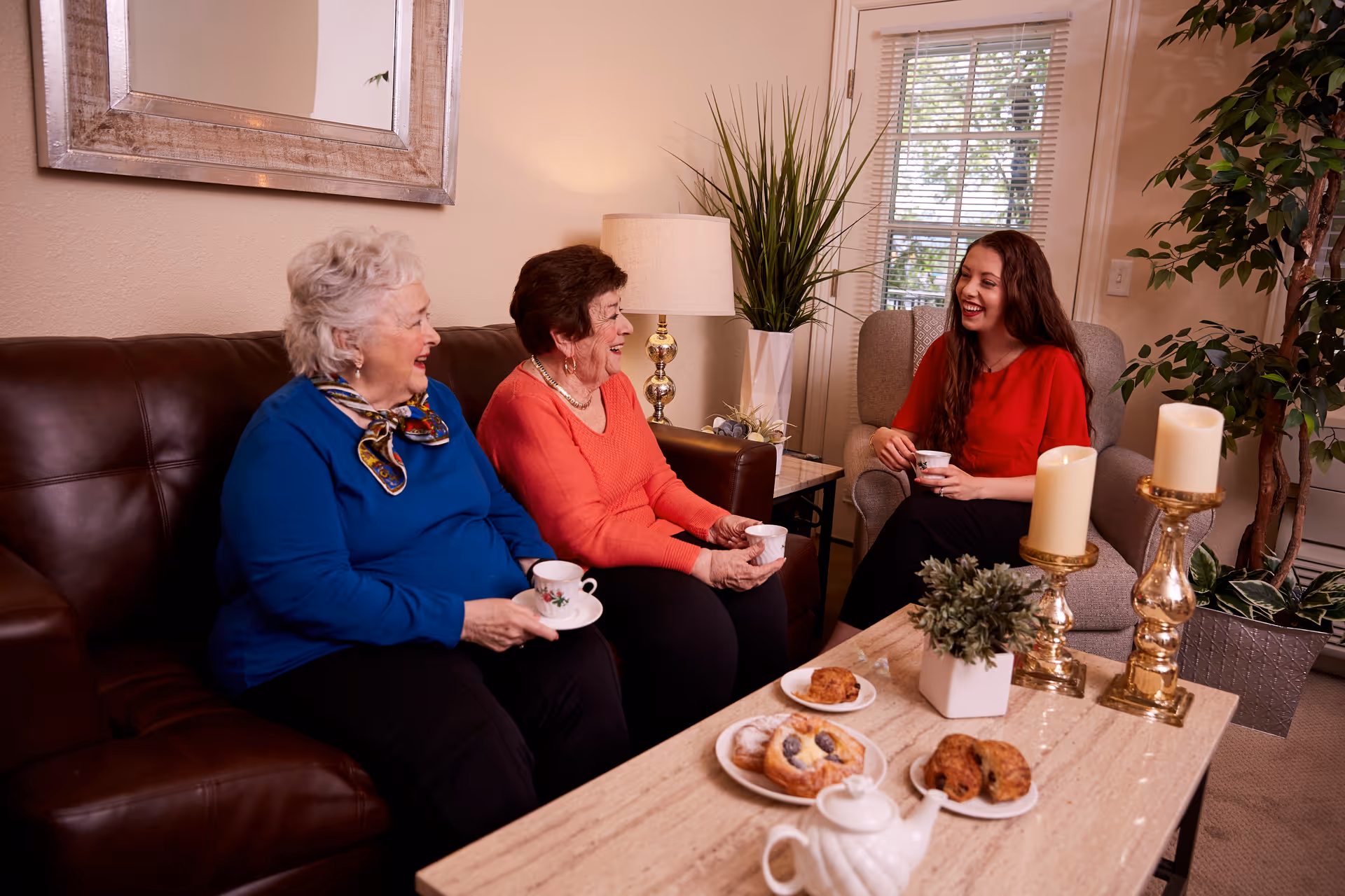 Three women sitting in a cozy living room, two elderly women on a brown leather couch and a younger woman on a gray armchair, all holding teacups and smiling. A coffee table in front of them has plates with pastries, a teapot, candles, and a small potted plant. The room has a large mirror on the wall, a lamp, and a window with blinds.