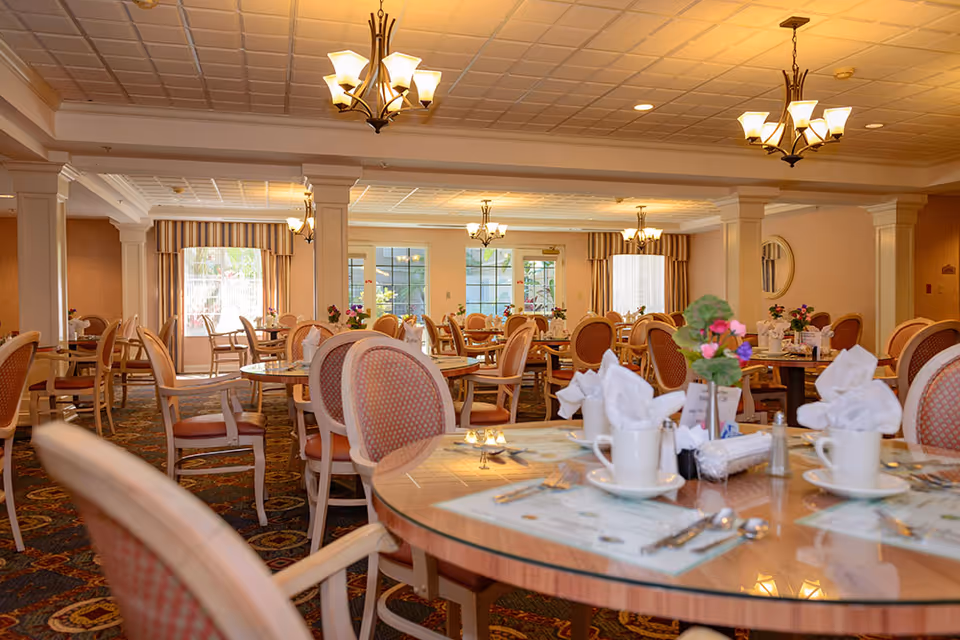 A spacious dining room with multiple round tables set with white napkins, cups, and silverware. The room features patterned carpet, beige walls, large windows with striped curtains, and several chandeliers hanging from a coffered ceiling. There are floral centerpieces on the tables.