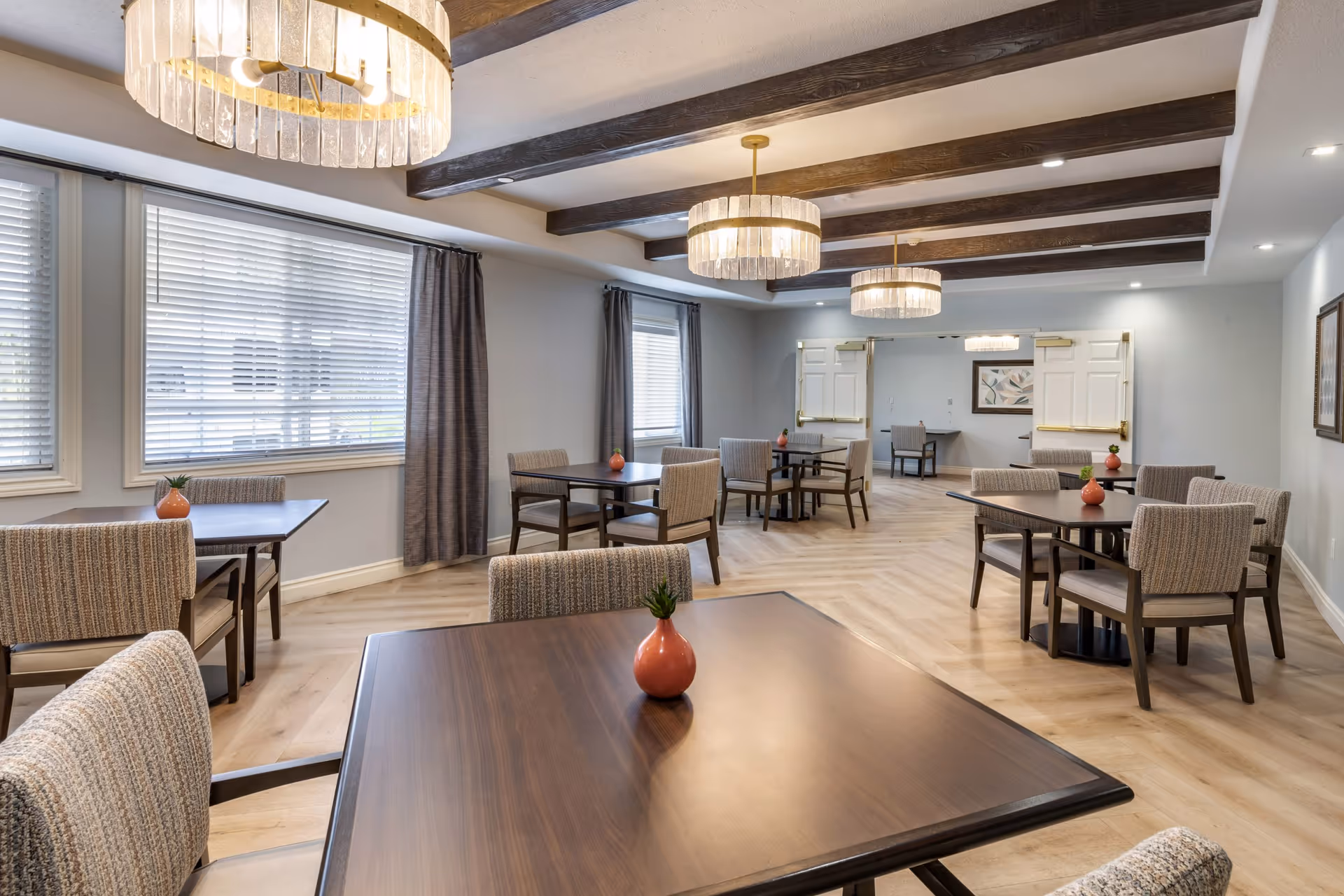 Bright dining room with multiple wooden tables and upholstered chairs, pendant chandeliers, exposed ceiling beams, and windows with blinds.