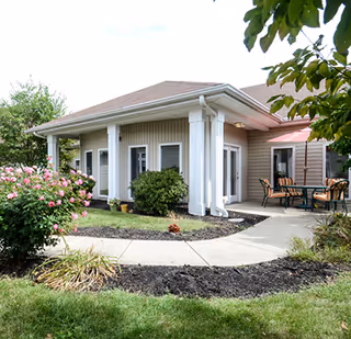 Exterior view of a single-story building with beige siding and white columns, surrounded by green grass, bushes, and flowering plants. There is a curved concrete walkway leading to a patio area with a table and chairs under a red umbrella.