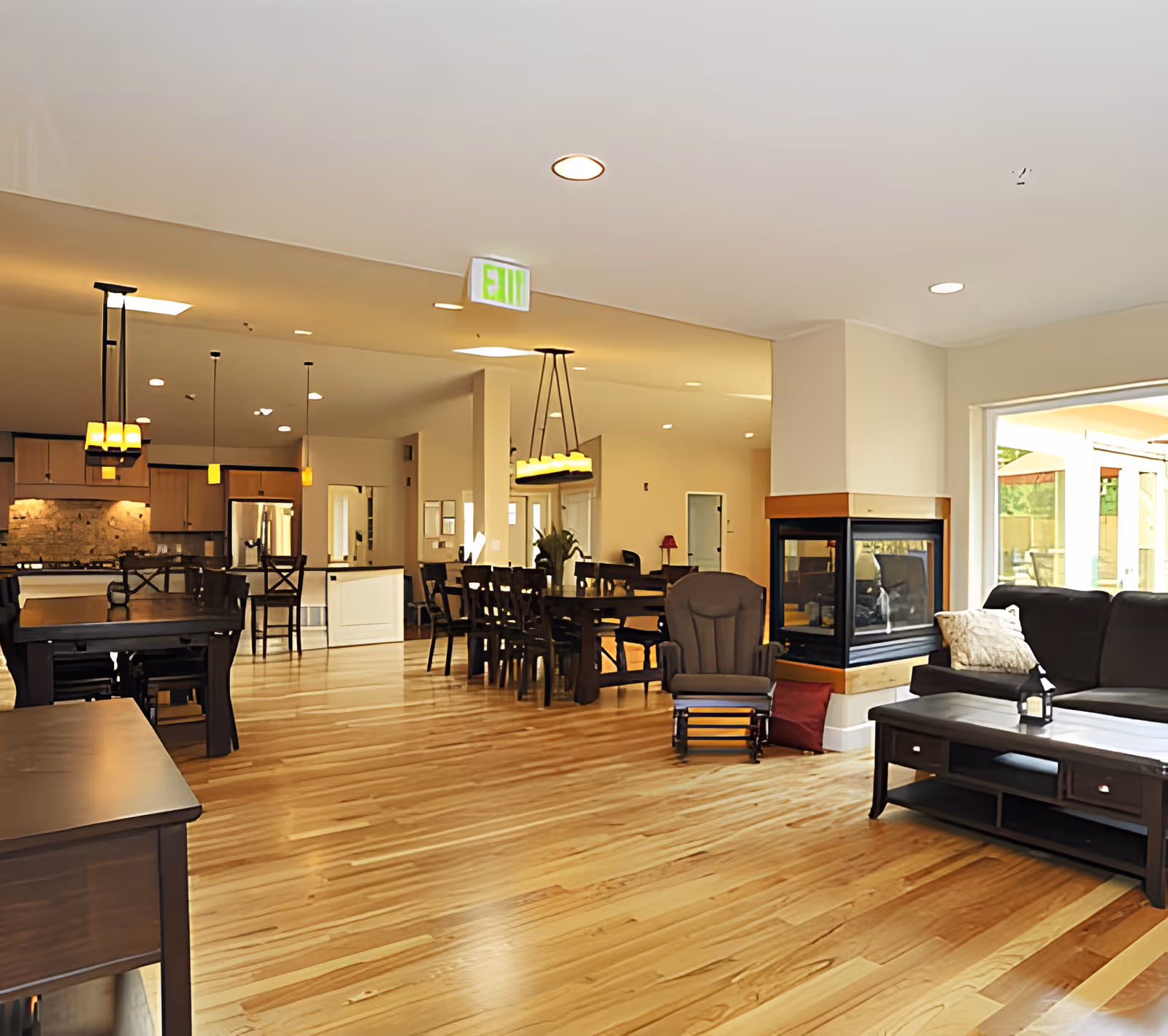 Spacious open-plan senior living common area with hardwood floors, seating and a fireplace, dining tables, and a kitchen in the background.
