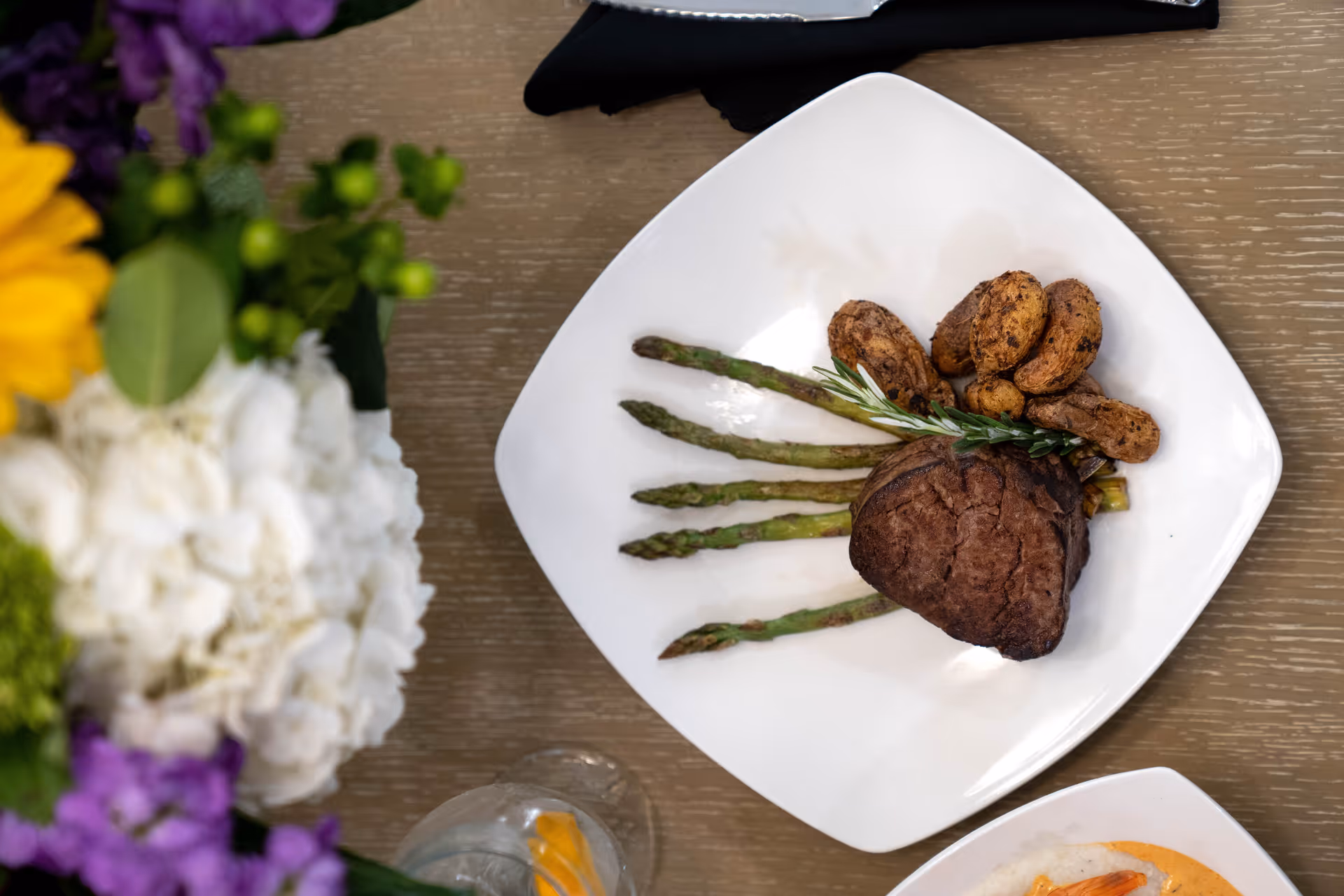 A white square plate with a serving of grilled asparagus, a piece of cooked steak garnished with a sprig of rosemary, and several roasted potatoes on a wooden table. There is a colorful flower arrangement and a glass of water with a lemon slice nearby.