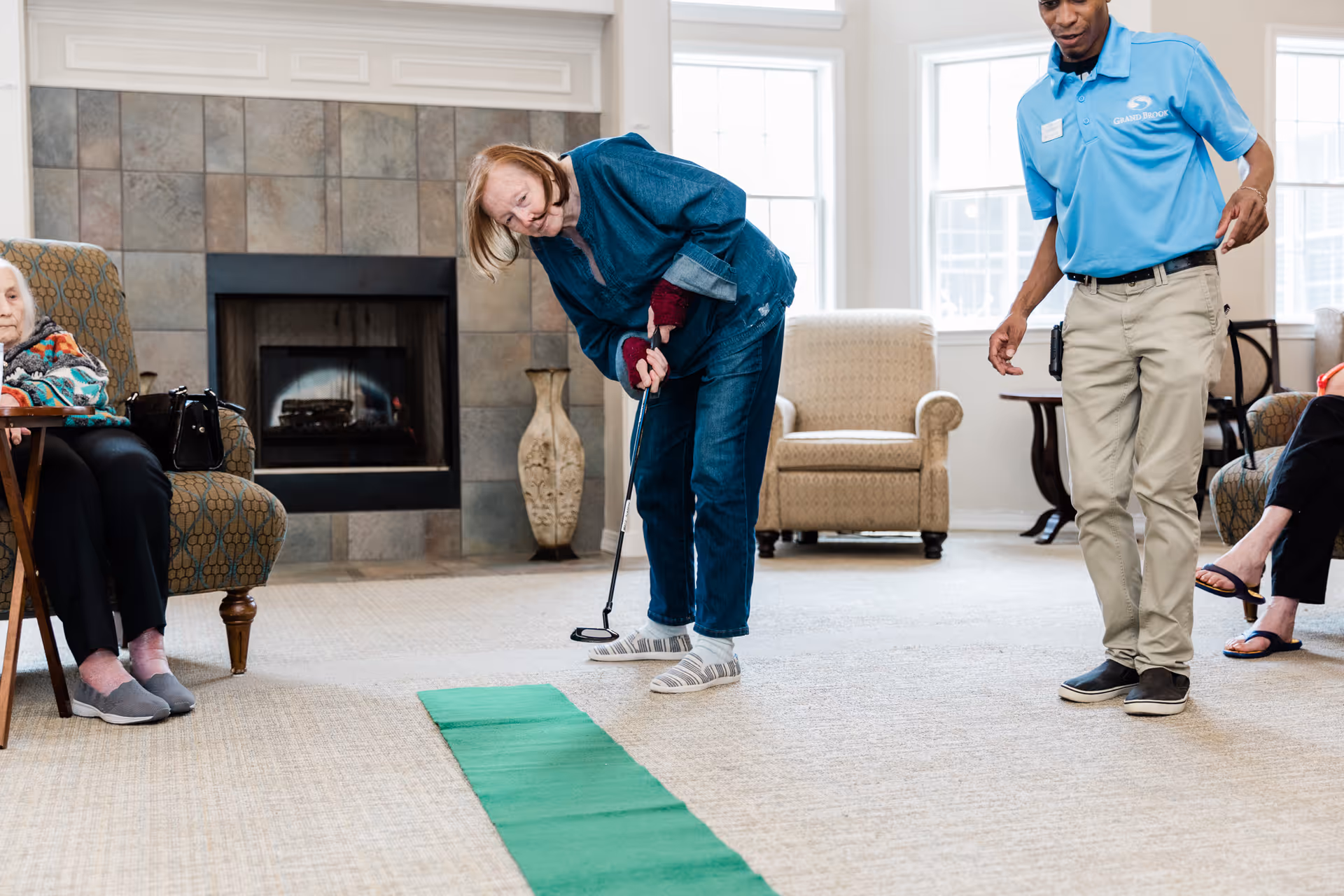 An elderly woman is playing indoor mini-golf on a green putting mat in a cozy living room area with a fireplace and armchairs. A staff member in a blue polo shirt and khaki pants watches and encourages her. Other elderly residents are seated nearby, observing the activity.