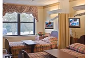 A bright hospital-style room with two beds separated by a privacy curtain. Each bed has a plaid blanket and a small overbed table. There are two windows with valance curtains letting in natural light, and a small table with a flower arrangement between the beds. Two wall-mounted televisions are visible above the beds.