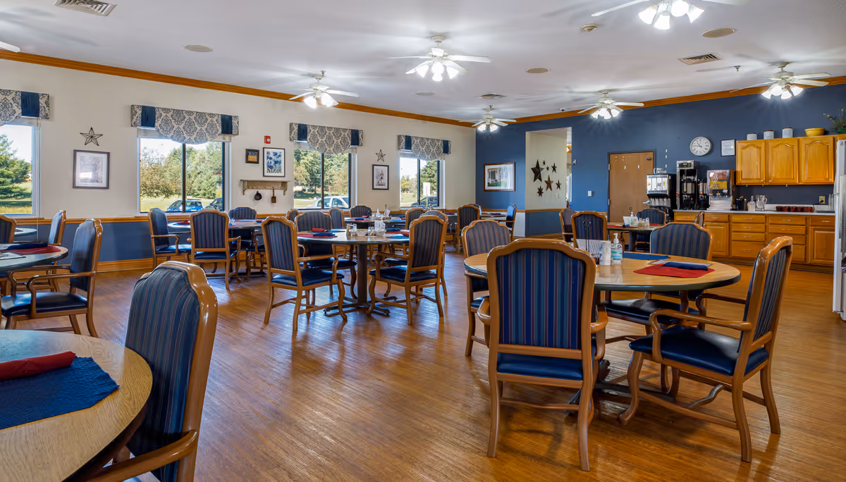 A spacious dining room with multiple round wooden tables surrounded by blue cushioned chairs. The room has large windows with patterned valances letting in natural light, ceiling fans with lights, and a wooden floor. In the background, there is a kitchenette area with wooden cabinets, a coffee station, and a clock on the wall.
