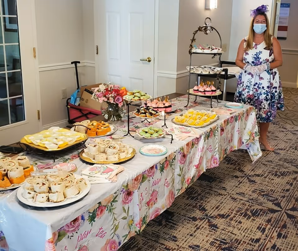 A woman wearing a floral dress, a purple fascinator, a face mask, and gloves stands next to a long table covered with a floral tablecloth. The table is set with various trays of pastries, cupcakes, and finger foods, along with a vase of flowers. The setting appears to be indoors with carpeted flooring and white walls.