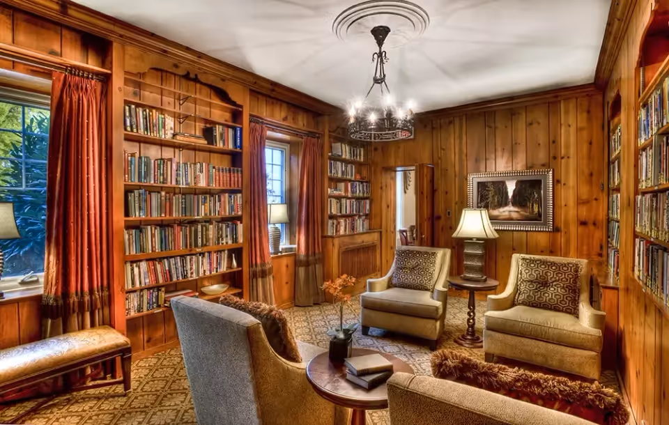 A cozy library room with wood-paneled walls, built-in bookshelves filled with books, two windows with red curtains, and a seating area consisting of four upholstered armchairs around a round wooden table with books and a small plant. A table lamp and a chandelier provide warm lighting, and a framed picture hangs on the wall.