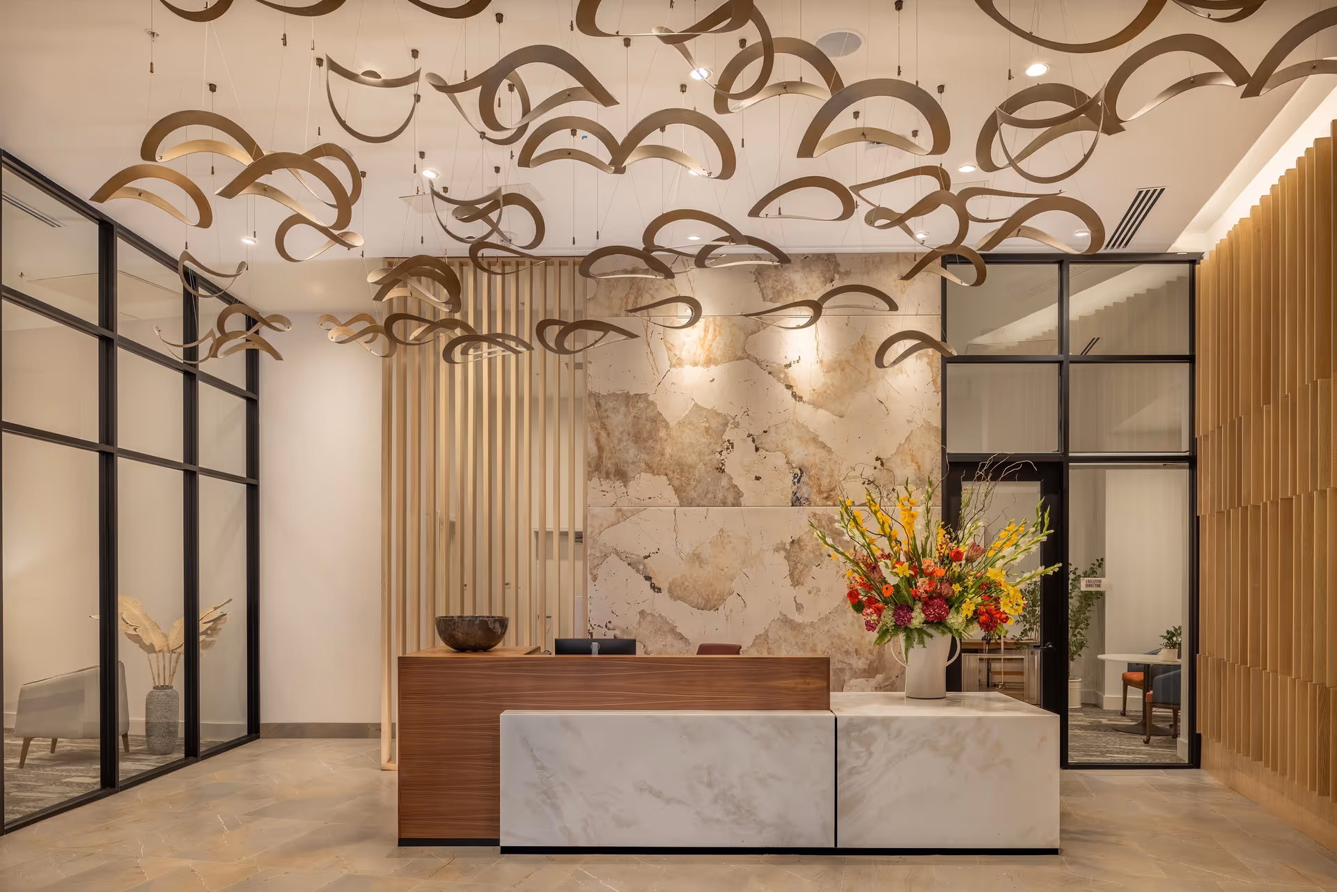 A modern reception area with a wooden and marble front desk, a large floral arrangement on the right side, decorative wooden ceiling art, and a stone accent wall behind the desk. Glass walls and doors are visible on the left and right sides, with a seating area seen through the glass on the left.